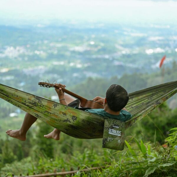Person enjoying a serene day in a hammock playing guitar with a scenic view in Indonesia.
