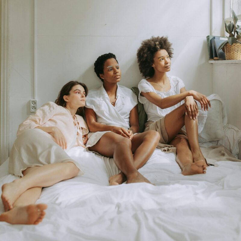 Three women of diverse backgrounds relaxing together on bed in cozy, sunlit room.